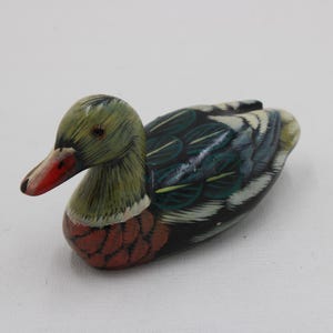 以下が含まれることがあります： 手描きの木製アヒルの置物。アヒルは緑色の頭、赤いクチバシ、黒、白、緑などの色の組み合わせの体を持っています。この置物は装飾品です。