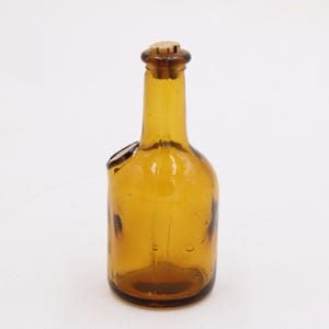 May include: Amber-colored glass bottle with a cork stopper. The bottle has a unique shape with a side opening. The glass has a slight texture and is transparent, allowing light to pass through. The bottle is likely vintage.