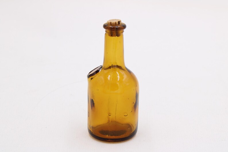 May include: Amber-colored glass bottle with a cork stopper. The bottle has a unique shape with a side opening. The glass has a slight texture and is transparent, allowing light to pass through. The bottle is likely vintage.