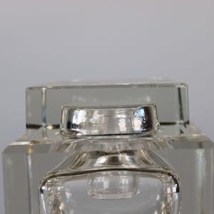 May include: A clear, square-shaped glass perfume bottle with a silver-colored cap. The bottle has a geometric design with a rectangular base and a square top. The cap is a shiny, metallic silver color.