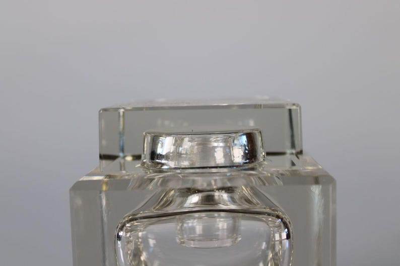 May include: A clear, square-shaped glass perfume bottle with a silver-colored cap. The bottle has a geometric design with a rectangular base and a square top. The cap is a shiny, metallic silver color.