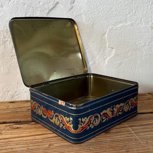 May include: An open, rectangular tin box with a navy blue exterior and a gold interior. The box is decorated with a red and gold ornate design along the edges. The box is sitting on a wooden surface.