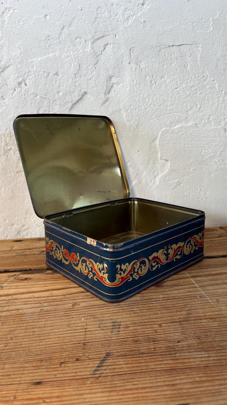 May include: An open, rectangular tin box with a navy blue exterior and a gold interior. The box is decorated with a red and gold ornate design along the edges. The box is sitting on a wooden surface.