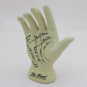 May include: A cream ceramic hand figurine, marked with black lines and text detailing palmistry features. The base is inscribed with "The Hand". This decorative item could be used by a fortune teller or spiritualist.
