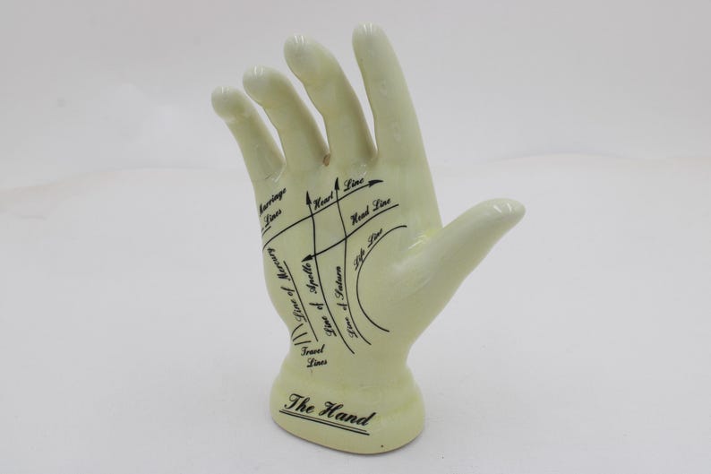 May include: A cream ceramic hand figurine, marked with black lines and text detailing palmistry features. The base is inscribed with "The Hand". This decorative item could be used by a fortune teller or spiritualist.