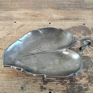 Peut inclure: Un plat argent&eacute; en forme de feuille avec une poign&eacute;e. Le plat pr&eacute;sente un motif de veines d&eacute;taill&eacute; et une poign&eacute;e l&eacute;g&egrave;rement incurv&eacute;e. La surface a un aspect us&eacute; et vintage, sugg&eacute;rant qu'il s'agit d'une pi&egrave;ce ancienne ou vintage.