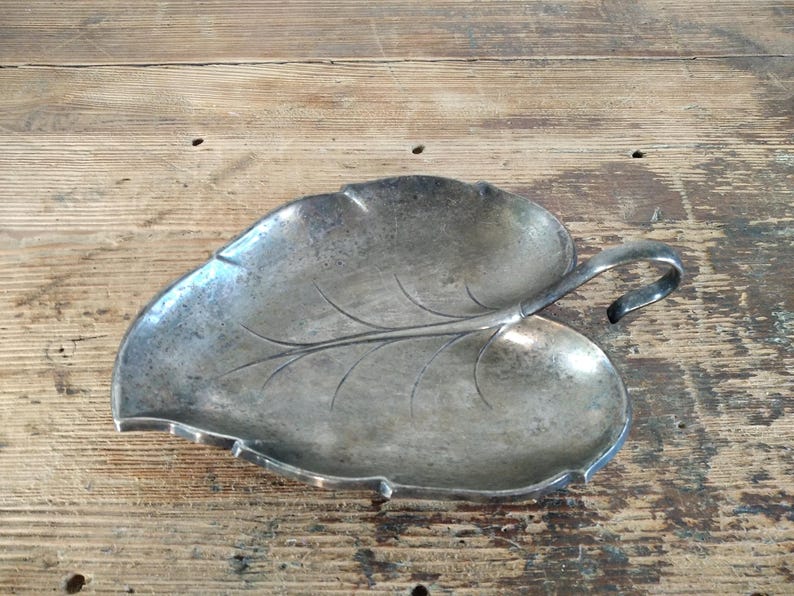 Peut inclure: Un plat argent&eacute; en forme de feuille avec une poign&eacute;e. Le plat pr&eacute;sente un motif de veines d&eacute;taill&eacute; et une poign&eacute;e l&eacute;g&egrave;rement incurv&eacute;e. La surface a un aspect us&eacute; et vintage, sugg&eacute;rant qu'il s'agit d'une pi&egrave;ce ancienne ou vintage.