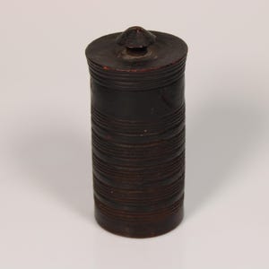 Puede incluir: Un recipiente cilíndrico de madera marrón oscuro con tapa. El recipiente tiene una serie de líneas horizontales talladas alrededor de su cuerpo. La tapa es redonda con un pequeño pomo cónico en la parte superior. Probablemente se utiliza para el almacenamiento.