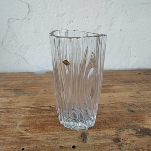 May include: A clear, textured glass vase with a unique, asymmetrical top. The vase features vertical, wave-like patterns and a small, gold-toned label. It stands on a wooden surface, showcasing its elegant design. The vase is approximately 10 inches tall.