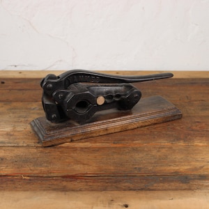 Op de afbeelding: Een vintage zwarte notenkraker met een lange hendel, rustend op een rechthoekige houten basis. De notenkraker is gemaakt van metaal en staat op een houten oppervlak. De achtergrond is een witte muur en een houten tafel.