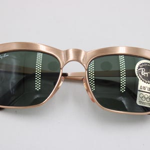 May include: Vintage Ray-Ban sunglasses with a gold-colored frame and dark green lenses. The sunglasses have a classic design with a curved brow bar and a keyhole bridge. A sticker on the lens indicates C-15 lens and the Ray-Ban brand.