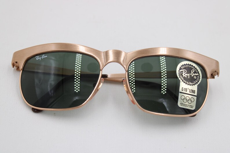 May include: Vintage Ray-Ban sunglasses with a gold-colored frame and dark green lenses. The sunglasses have a classic design with a curved brow bar and a keyhole bridge. A sticker on the lens indicates C-15 lens and the Ray-Ban brand.