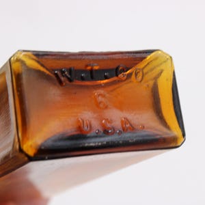 May include: Close-up of the bottom of a rectangular amber glass bottle. The base has embossed text that reads "W.T. & CO." above the number "6" and "U.S.A." below. The bottle has a vintage appearance.