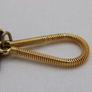 May include: Close-up of a gold-colored metal key ring with a coiled spring design. The key ring is attached to a rectangular burgundy leather tag with gold trim. The background is a plain white surface.