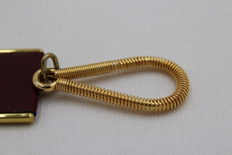 May include: Close-up of a gold-colored metal key ring with a coiled spring design. The key ring is attached to a rectangular burgundy leather tag with gold trim. The background is a plain white surface.