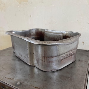 Puede incluir: Un recipiente rectangular de metal desgastado con la palabra "Polybox" grabada en el lateral. El recipiente tiene una forma ligeramente curvada y una extensión en forma de asa en un lado. El metal tiene un aspecto desgastado y envejecido.