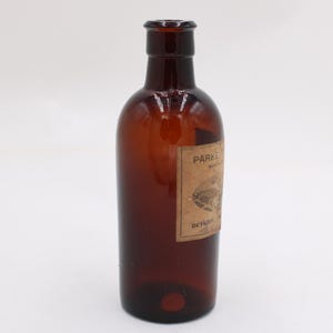 Puede incluir: Una botella de vidrio marr&oacute;n con una etiqueta. La botella tiene un cuerpo redondeado y un cuello estrecho. La etiqueta es rectangular y tiene texto y una ilustraci&oacute;n. La botella est&aacute; vac&iacute;a y parece ser vintage.