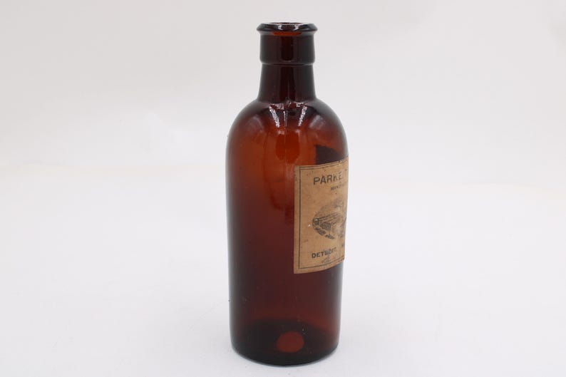 Puede incluir: Una botella de vidrio marr&oacute;n con una etiqueta. La botella tiene un cuerpo redondeado y un cuello estrecho. La etiqueta es rectangular y tiene texto y una ilustraci&oacute;n. La botella est&aacute; vac&iacute;a y parece ser vintage.