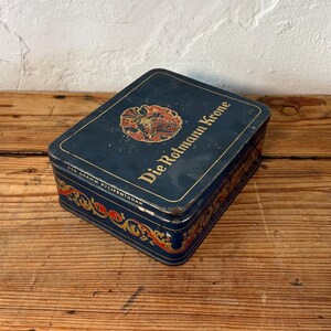 May include: A rectangular, vintage blue tin with the text "Die Rotmann Krone" and decorative gold and red accents. The tin also has the text "250 Gramm Pfeifentabak".