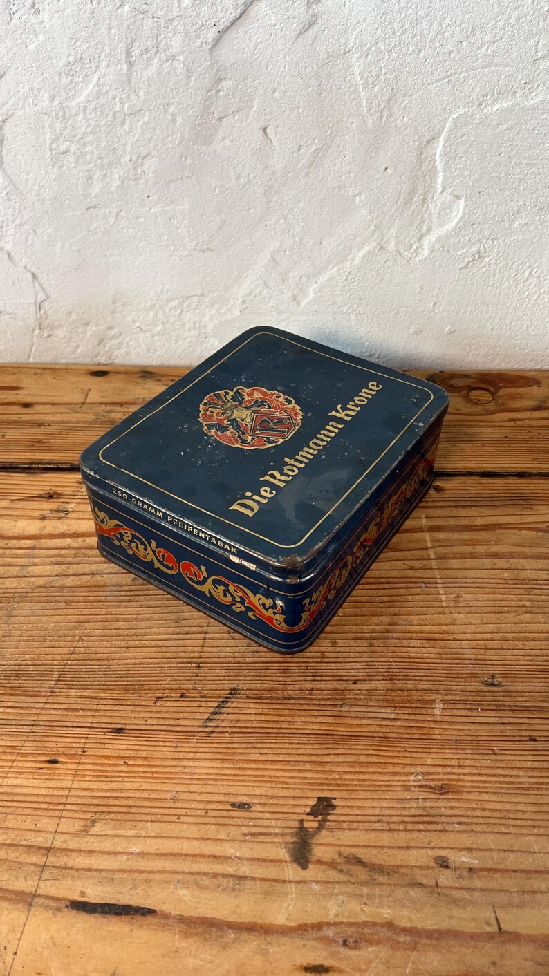 May include: A rectangular, vintage blue tin with the text "Die Rotmann Krone" and decorative gold and red accents. The tin also has the text "250 Gramm Pfeifentabak".