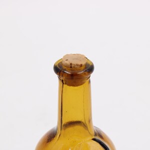 May include: A close-up of a vintage amber glass bottle with a cork stopper. The bottle has a rounded base and a long, slender neck. The cork is a light brown color, and the glass has a slight texture.