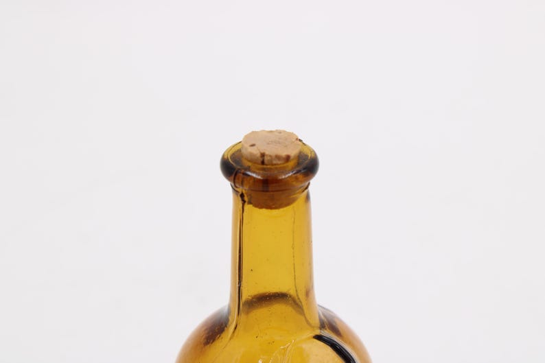May include: A close-up of a vintage amber glass bottle with a cork stopper. The bottle has a rounded base and a long, slender neck. The cork is a light brown color, and the glass has a slight texture.