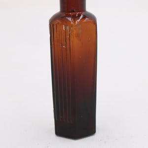 Puede incluir: Una botella de vidrio marr&oacute;n vintage con cuerpo rectangular y cuello estrecho. La botella tiene estr&iacute;as verticales en la parte delantera y los lados. El vidrio es de color &aacute;mbar oscuro. La botella est&aacute; vac&iacute;a.