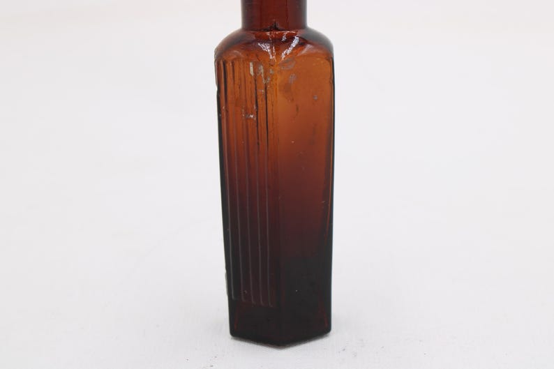 Puede incluir: Una botella de vidrio marr&oacute;n vintage con cuerpo rectangular y cuello estrecho. La botella tiene estr&iacute;as verticales en la parte delantera y los lados. El vidrio es de color &aacute;mbar oscuro. La botella est&aacute; vac&iacute;a.