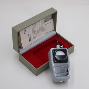 Könnte beinhalten: Ein Vintage-Gerät in Grau und Schwarz mit Messgerät und Zifferblatt, neben einer offenen grauen Box mit rotem Innenraum. Die Box enthält Papiere. Das Gerät trägt den Text "Neurex".