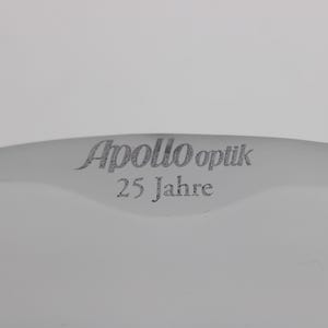 K&ouml;nnte beinhalten: Ein gebogenes, wei&szlig;es Objekt mit dem Text "Apollo Optik 25 Jahre" in grauer, stilisierter Schrift. Der Text ist auf dem Objekt zentriert, das Teil eines gr&ouml;&szlig;eren Gegenstandes zu sein scheint.