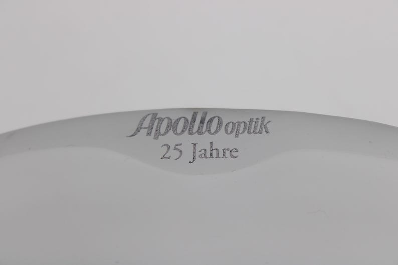 K&ouml;nnte beinhalten: Ein gebogenes, wei&szlig;es Objekt mit dem Text "Apollo Optik 25 Jahre" in grauer, stilisierter Schrift. Der Text ist auf dem Objekt zentriert, das Teil eines gr&ouml;&szlig;eren Gegenstandes zu sein scheint.