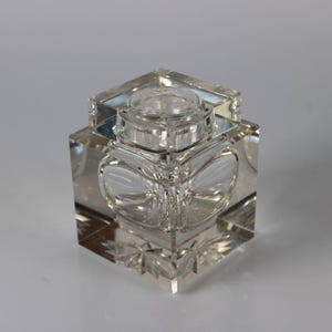 May include: A clear, square-shaped glass inkwell with a lid. The inkwell features a decorative, etched design on the sides and base. The lid is round and sits atop the square body. The inkwell is displayed on a white surface.