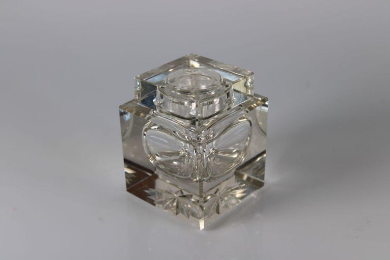 May include: A clear, square-shaped glass inkwell with a lid. The inkwell features a decorative, etched design on the sides and base. The lid is round and sits atop the square body. The inkwell is displayed on a white surface.