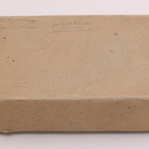 May include: A rectangular, closed cardboard box in a light brown color. The box has text printed on the top, including the name "Herbert Kaiser". The box appears to be sealed with staples on the edges.