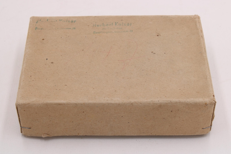 May include: A rectangular, closed cardboard box in a light brown color. The box has text printed on the top, including the name "Herbert Kaiser". The box appears to be sealed with staples on the edges.