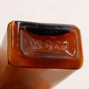 May include: Close-up of a brown glass bottle with a rectangular shape. The bottom of the bottle has the embossed text "L W G Co". The bottle has a dark brown top and a lighter brown body.