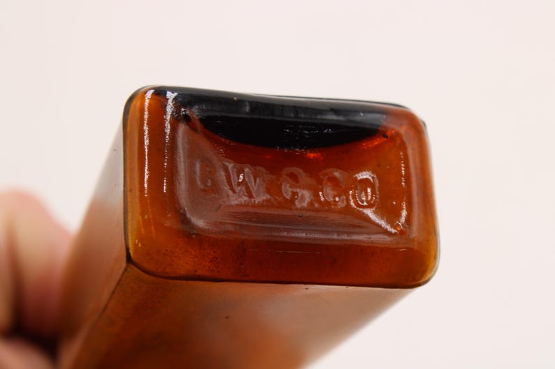May include: Close-up of a brown glass bottle with a rectangular shape. The bottom of the bottle has the embossed text "L W G Co". The bottle has a dark brown top and a lighter brown body.