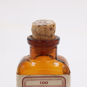 May include: Amber-colored glass bottle with a cork stopper. The bottle has a brown neck and a label with the number "100". The cork is light brown and has a circular marking on top. The bottle is likely for storing liquids.