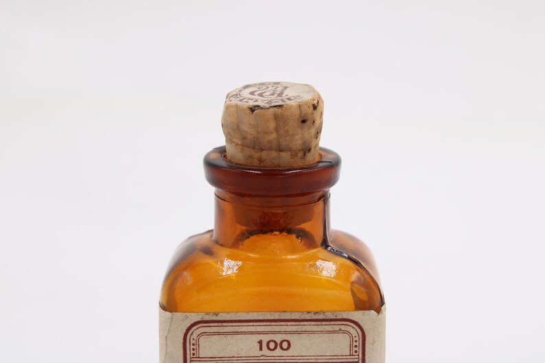 May include: Amber-colored glass bottle with a cork stopper. The bottle has a brown neck and a label with the number "100". The cork is light brown and has a circular marking on top. The bottle is likely for storing liquids.
