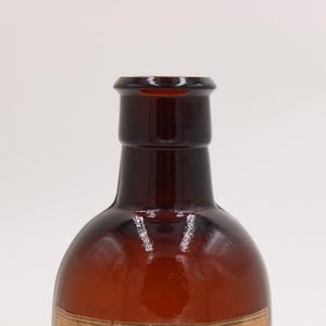 Puede incluir: Primer plano de una botella de vidrio marr&oacute;n con un cuerpo redondeado y un cuello estrecho. La botella tiene un color &aacute;mbar oscuro y una superficie lisa y brillante. La botella est&aacute; vac&iacute;a y parece ser vintage o antigua.
