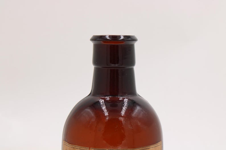 Puede incluir: Primer plano de una botella de vidrio marr&oacute;n con un cuerpo redondeado y un cuello estrecho. La botella tiene un color &aacute;mbar oscuro y una superficie lisa y brillante. La botella est&aacute; vac&iacute;a y parece ser vintage o antigua.
