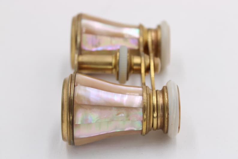 May include: Antique opera glasses with a gold-tone metal frame and iridescent mother-of-pearl inlay. The lenses are white, and the binoculars are connected by a gold-tone bridge. A classic accessory.