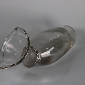 May include: A clear glass decanter with a wide, flared spout. The decanter is lying on its side, showcasing its elegant shape and design. The glass appears to be smooth and unadorned, reflecting the light.