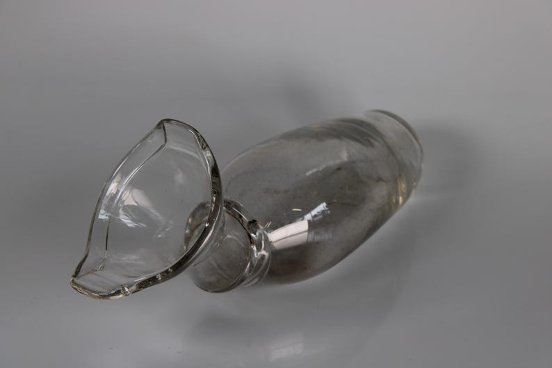 May include: A clear glass decanter with a wide, flared spout. The decanter is lying on its side, showcasing its elegant shape and design. The glass appears to be smooth and unadorned, reflecting the light.