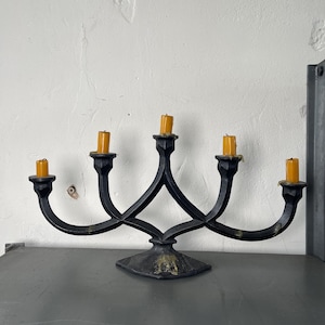 May include: A black wrought iron candelabra with five candle holders, each holding a yellow candle. The candelabra has a decorative base and curved arms. The background is a white wall.