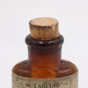 May include: Close-up of a vintage brown glass bottle with a cork stopper. The bottle has a rounded shape and a label around the base. The cork is light brown and the glass has a gradient from dark brown to clear.