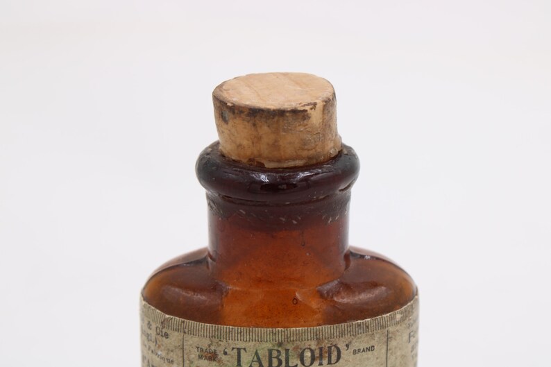 May include: Close-up of a vintage brown glass bottle with a cork stopper. The bottle has a rounded shape and a label around the base. The cork is light brown and the glass has a gradient from dark brown to clear.