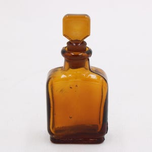 May include: Amber glass bottle with a square base and a matching stopper. The bottle has a rounded shoulder and a rectangular stopper. The glass has a slight amber hue, and the bottle is empty.