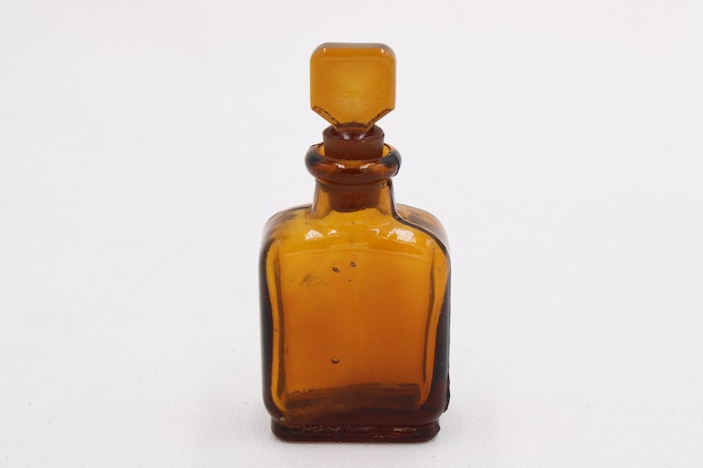 May include: Amber glass bottle with a square base and a matching stopper. The bottle has a rounded shoulder and a rectangular stopper. The glass has a slight amber hue, and the bottle is empty.