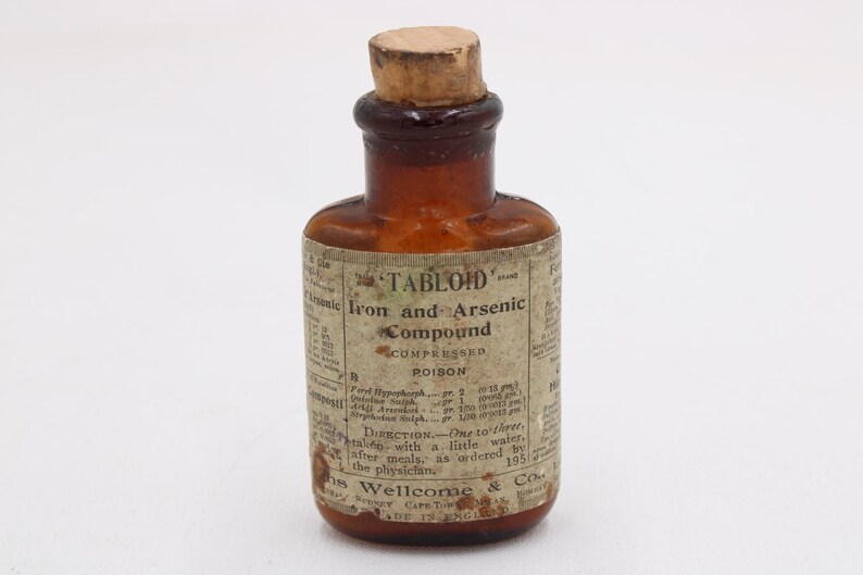 May include: A small, antique brown glass bottle with a cork stopper. The label reads "TABLOID" and "Iron and Arsenic Compound, Compressed Poison." The bottle has a vintage aesthetic, with faded text and a classic design.